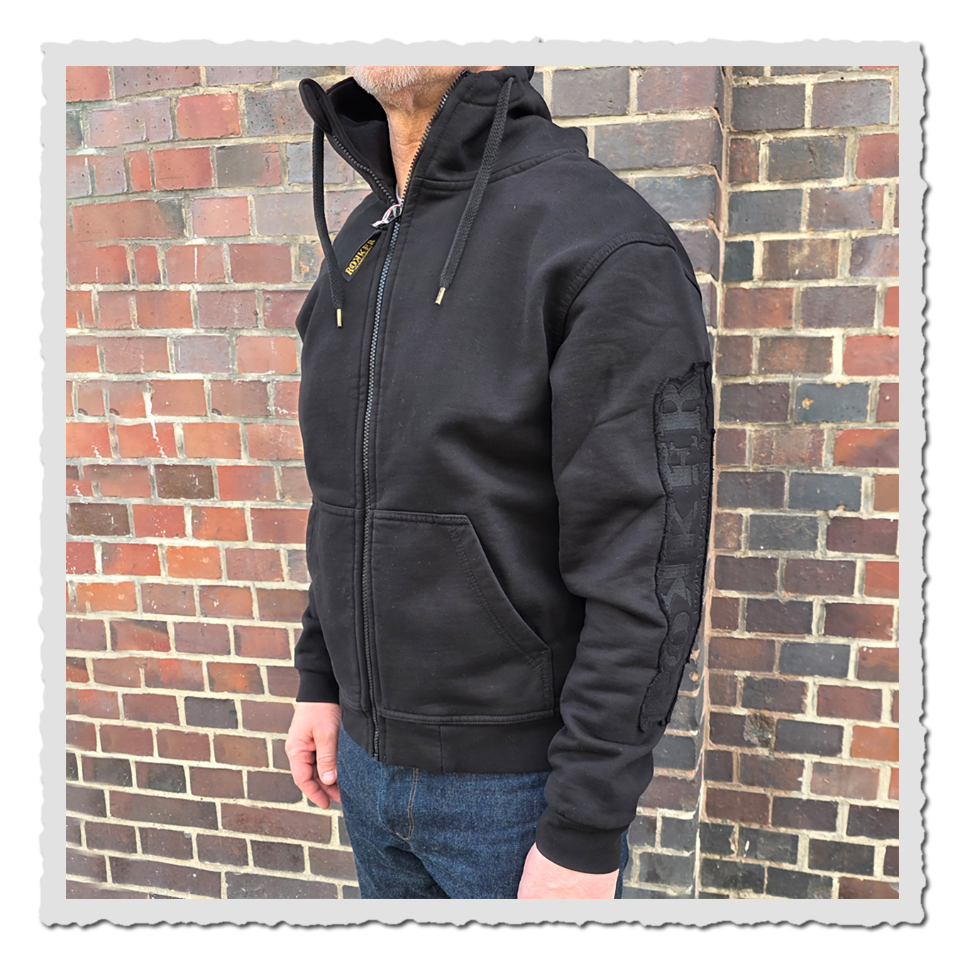 Seitliche Ansicht einer Person mit schwarzem Kapuzenhoodie vor gemauerter Backsteinwand im Außenbereich.